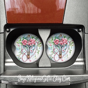 May include: Two car coasters with a watercolor design featuring a cross, pink roses, and a beaded rosary. The coasters are round and fit into a car's cup holders. The background is a light blue and white watercolor wash.