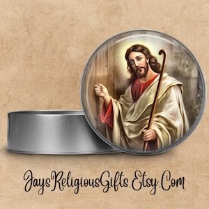 May include: A round, silver-colored metal container with a glass-covered image of Jesus holding a staff. The image features Jesus with long brown hair and a beard, wearing a white robe and red undergarment. The text "JaysReligiousGifts.Etsy.Com" is at the bottom.