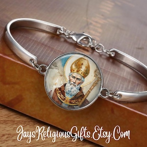 May include: Silver-toned bracelet featuring a round charm with a religious image of a man with a white beard, wearing a gold crown and holding a staff. The charm is set against a light blue and white background.