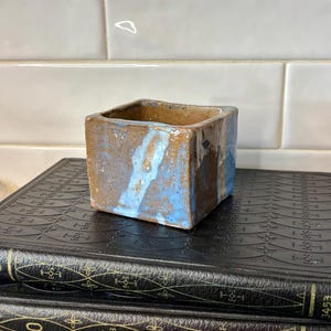 May include: A small, square ceramic planter with a textured brown and blue design. The planter sits atop a stack of three antique books with black covers and gold accents. The background features white rectangular tiles.