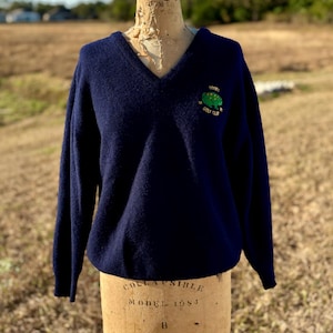 May include: A navy blue V-neck sweater with long sleeves. The sweater has an embroidered green tree logo with the text "Dunes Golf Club". The sweater is displayed on a vintage mannequin.
