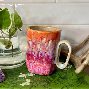 May include: A handmade ceramic mug with a textured surface and a unique handle. The mug features a gradient of colors, transitioning from orange to pink and purple. The mug is sitting on a green book with the text "HOUSEPLANTS & INDOOR GARDENING".