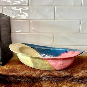 May include: A handmade ceramic bowl with a wide, shallow design. The bowl features a color-blocked pattern with sections of blue, pink, yellow, and green. The bowl is displayed on a brown surface with two antique books in the background.