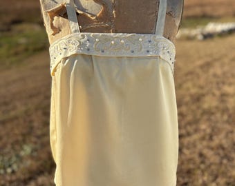 May include: A pale yellow top with thin straps and a decorative band across the chest. The band is embellished with small pearls and beads. The top is displayed on a vintage dress form.