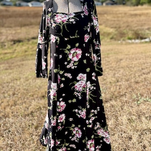 May include: A black floral print dress with long sleeves and a square neckline. The dress features a pattern of pink and white flowers with green leaves. The dress is displayed on a vintage dress form against a backdrop of a field and blue sky.
