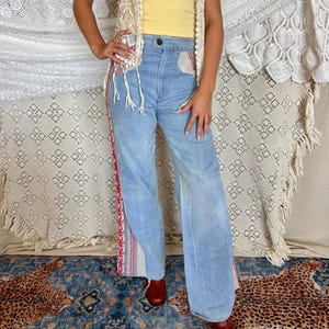 May include: A yellow tube top paired with a cream-colored macrame vest featuring fringe trim and light blue denim jeans with a red and white patterned side stripe detail and a single dark button closure at the waistline.
