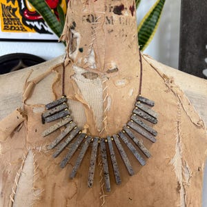 May include: A necklace featuring a series of rectangular, speckled grey stone beads arranged in a fan-like pattern. The beads are separated by small, round, metallic gold-toned spacers and are strung on a thin, dark brown cord.