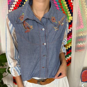 May include: A blue chambray button-down shirt featuring embroidered bird and floral motifs on the collar area, long sleeves with beige and blue vertical stripes, a pointed collar, and a front button closure with visible stitching details.