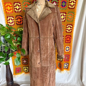 May include: A long, brown suede coat featuring a tan faux fur lining, a wide notched collar, and three front buttons. The coat has visible seam detailing along the front panels and sleeves, with faux fur trim visible at the cuffs and collar.