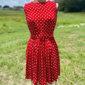 May include: A sleeveless red dress featuring a white polka dot pattern throughout. The dress includes a matching red fabric belt with a rectangular buckle at the waist and a pleated skirt that falls above the knee, displayed on a dress form.