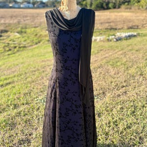May include: A sleeveless black dress with a floral pattern and a draped cowl neckline. The dress is displayed on a vintage dress form outdoors, with a long black scarf draped over the shoulder. The dress has a flowing skirt.
