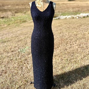 May include: A navy blue, sleeveless, floor-length dress with a V-neckline. The dress is covered in small, sparkling embellishments, giving it a textured appearance. The dress is displayed on a vintage dress form outdoors.