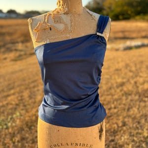 May include: A navy blue one-shoulder top with a decorative rhinestone clasp. The top is displayed on a vintage dress form. The dress form has the words "COLLAPSIBLE MODEL 1954" printed on it.