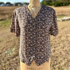 May include: A vintage floral print blouse with short sleeves and a V-neck. The blouse features a dark background with a pattern of small red, white, and blue flowers. It is displayed on a vintage dress form.