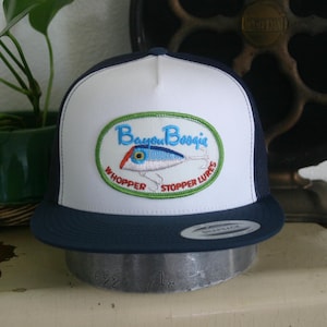 May include: A white and navy blue trucker hat with a green, white, and red embroidered patch that says "Bayou Boogie" and "Whopper Stopper Lures".