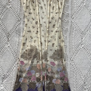 May include: A sheer, open-front duster in cream with a floral and tree print. The bottom hem features a purple border with a floral design in pink, blue, and peach. The duster is sleeveless and has a relaxed fit.