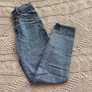 May include: Light blue denim jeans with a high waist and button fly closure. The jeans are slightly faded and have a classic five-pocket design. The pants are folded on a textured, light brown surface.