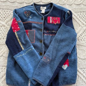 May include: A blue patchwork cardigan with various decorative patches. The cardigan features a heart and floral embroidery, along with button closures. The sleeves and body are a mix of denim and other blue fabrics.