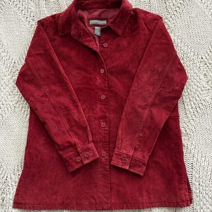 May include: A red suede button-down shirt with long sleeves. The shirt has a collared neckline and buttoned cuffs. The fabric appears soft and textured. The shirt is laid flat on a cream-colored, textured surface.