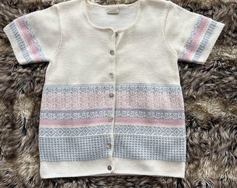 Può includere: Cardigan color crema a maniche corte con scollo tondo e chiusura con bottoni. Le maniche hanno strisce rosa e blu. Il corpo presenta fasce orizzontali con disegni rosa e blu.