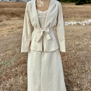 May include: A cream-colored vintage skirt suit displayed on a mannequin. The suit includes a long-sleeved jacket with a V-neck, a matching skirt, and a top with a ruched bodice. A cream-colored bow cinches the waist.