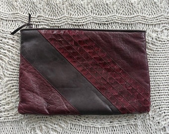 May include: A rectangular clutch with a burgundy and dark brown color scheme. The burgundy section has a textured, quilted pattern. A dark brown diagonal stripe bisects the burgundy section. A black zipper pull is visible.