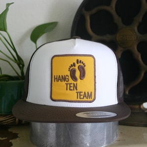 May include: A white and brown trucker hat with a yellow square patch that says "Hang Ten Team" with a brown footprint graphic.