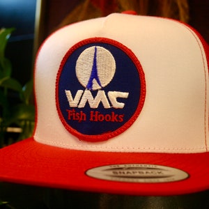 May include: A red and white trucker hat with a red brim. The hat features a circular patch with a blue background, a white Eiffel Tower design, and the text "VMC Fish Hooks" in white and red.