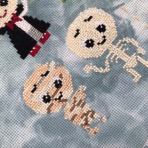 May include: Cross-stitch design featuring three Halloween characters: a vampire, a skeleton, and a mummy. The vampire wears a black cape with a white shirt and red accents. The skeleton and mummy are beige with black details.