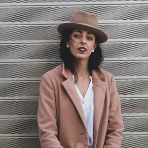 Op de afbeelding: Een vrouw draagt een bruine fedora, een lichtbruine jas en een wit shirt. Ze staat voor een metalen muur met een patroon van kleine gaten.
