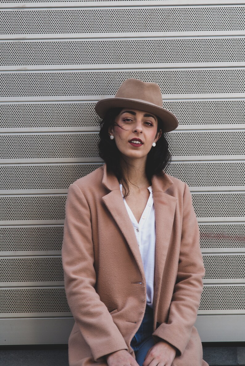 Op de afbeelding: Een vrouw draagt een bruine fedora, een lichtbruine jas en een wit shirt. Ze staat voor een metalen muur met een patroon van kleine gaten.