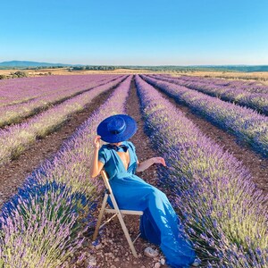May include: A woman wearing a blue dress and a blue hat sits on a wooden chair in a field of lavender.