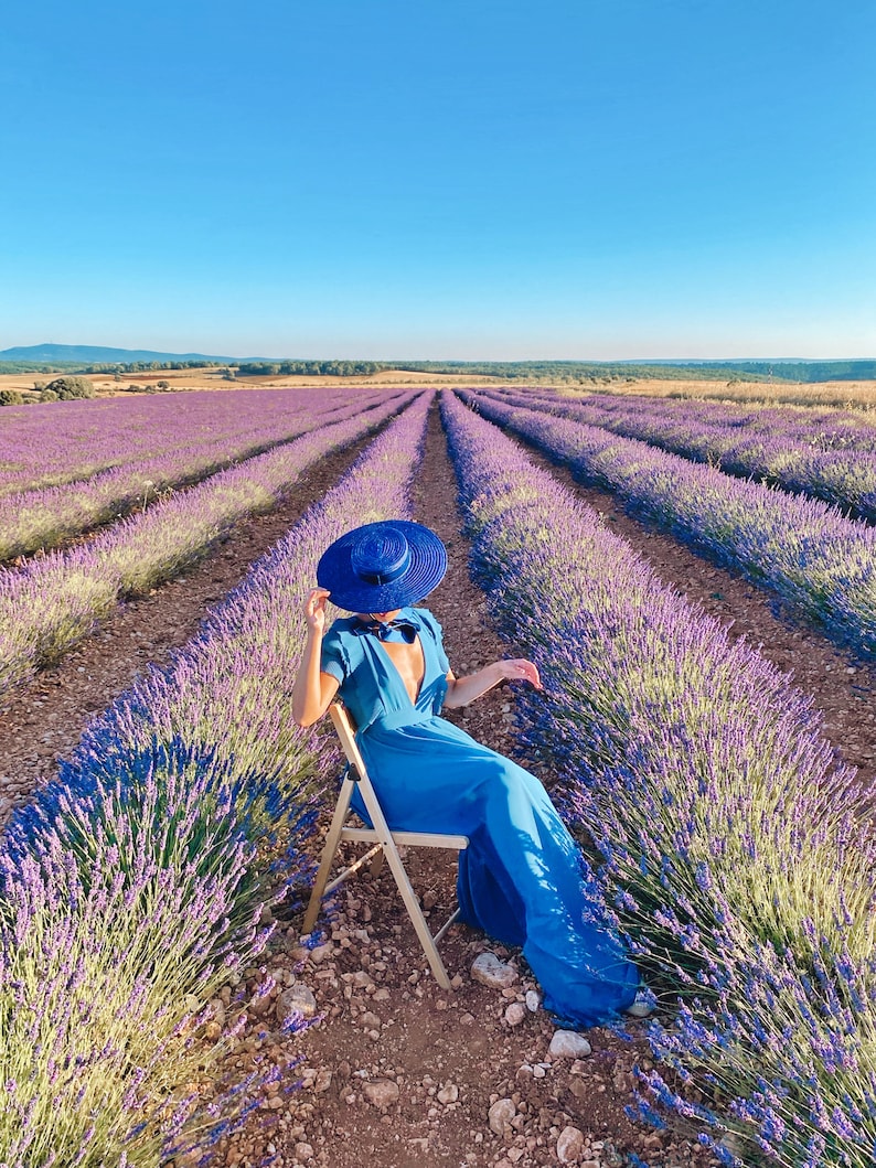 May include: A woman wearing a blue dress and a blue hat sits on a wooden chair in a field of lavender.