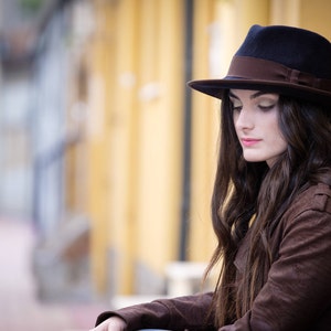 Op de afbeelding: Een vrouw met lang bruin haar die een bruine su&egrave;de jas en een zwarte fedora met een bruine band draagt.
