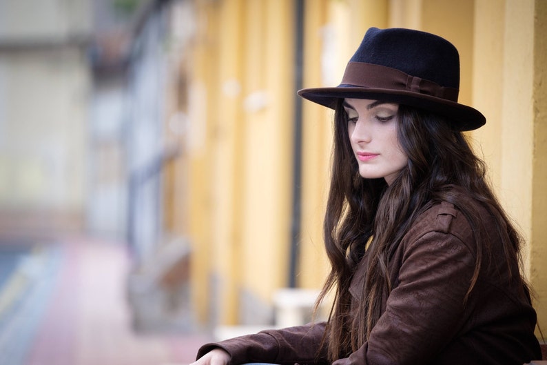 Op de afbeelding: Een vrouw met lang bruin haar die een bruine su&egrave;de jas en een zwarte fedora met een bruine band draagt.