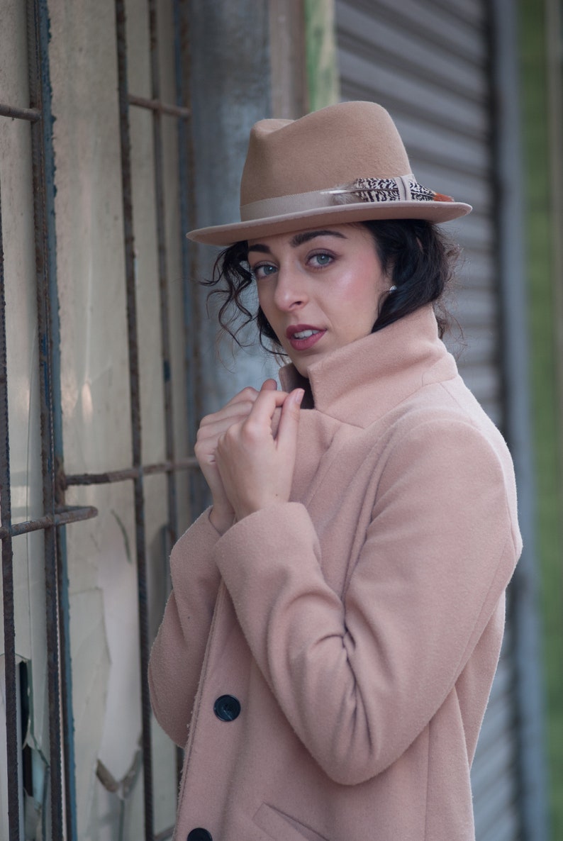 Op de afbeelding: Een vrouw draagt een lichtbruine jas met een bijpassende fedora-hoed. De hoed heeft een bruine verenversiering. De vrouw staat voor een metalen rooster.