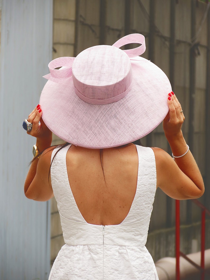 May include: A light pink, wide-brimmed hat with a matching ribbon bow. The hat has a rounded crown and a broad brim. The person is wearing a white dress with a low back. The person is wearing a gold watch and a silver bracelet.