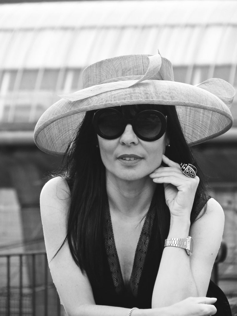 May include: A woman wearing a wide-brimmed hat, large sunglasses, and a black dress. The hat is light-colored with a decorative bow. She is wearing a silver watch and a large ring. The image is in black and white.