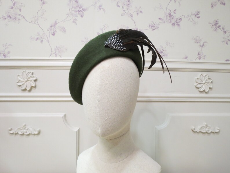 Op de afbeelding: Een donkergroene baret met een zwarte veer en een zilveren en zwarte broche. De baret zit op een wit mannequin hoofd.