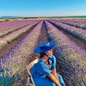 May include: A woman wearing a blue dress and a blue hat sits on a wooden chair in a field of lavender.