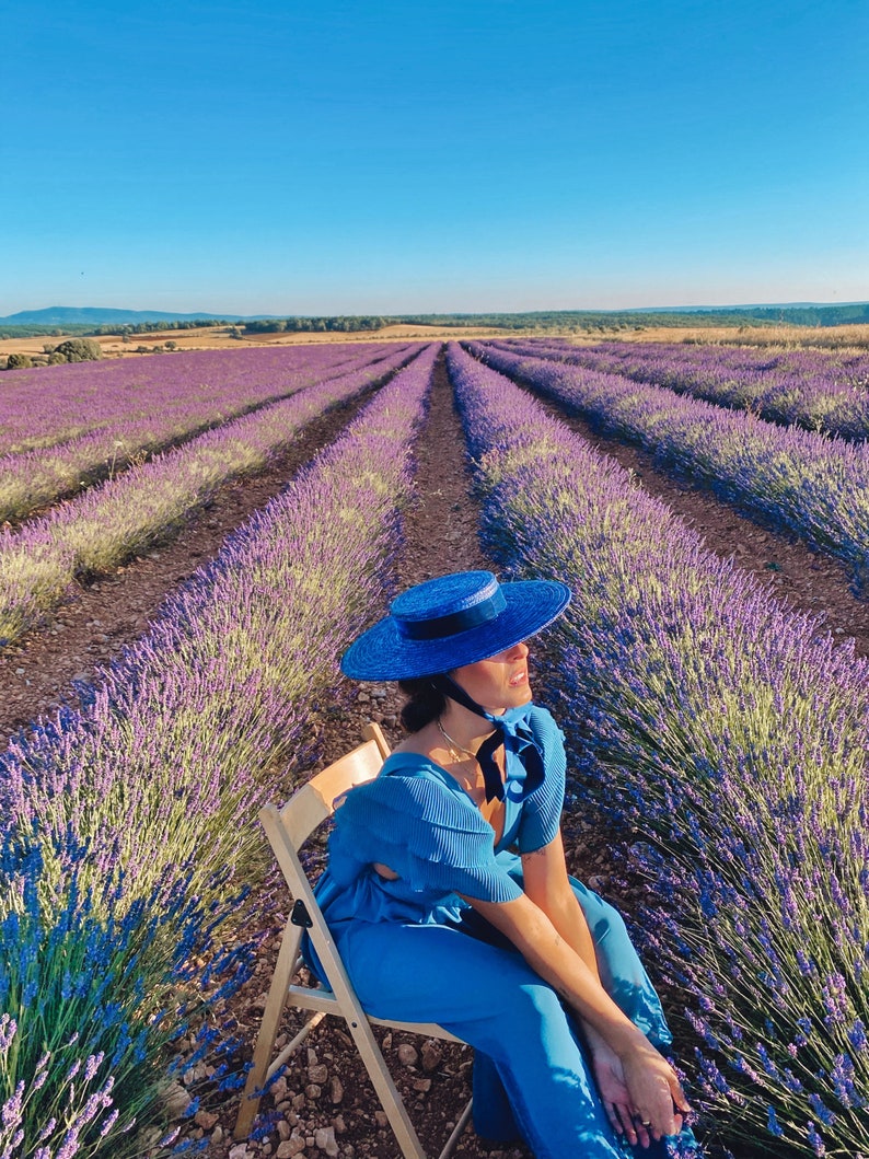 May include: A woman wearing a blue dress and a blue hat sits on a wooden chair in a field of lavender.