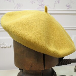 May include: A yellow wool beret with a small pompom on top. The beret is sitting on a wooden hat form.
