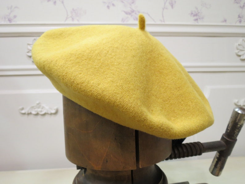 May include: A yellow wool beret with a small pompom on top. The beret is sitting on a wooden hat form.