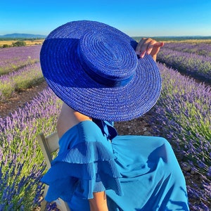 May include: A woman wearing a blue dress and a large blue sun hat sits in a chair in a field of lavender.