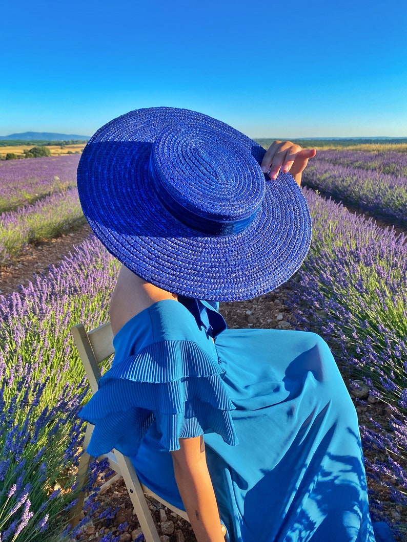 May include: A woman wearing a blue dress and a large blue sun hat sits in a chair in a field of lavender.