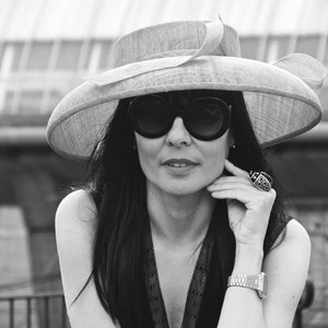 May include: A woman wearing a wide-brimmed hat, large sunglasses, and a black dress. The hat is light-colored with a decorative bow. She is wearing a large ring and a silver watch. The image is in black and white.