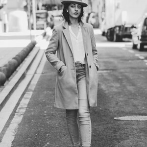 Op de afbeelding: Een vrouw in een bruine fedora, een beige jas en een blauwe jeans loopt over een straat in de stad.