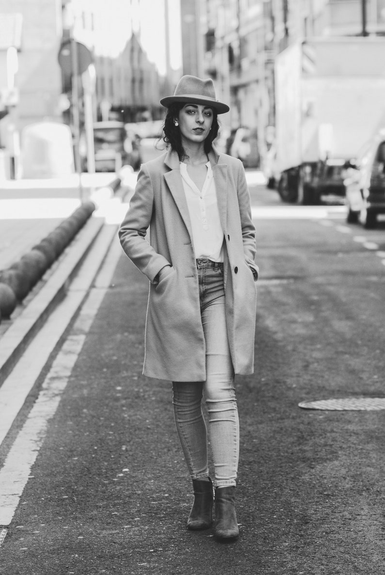 Op de afbeelding: Een vrouw in een bruine fedora, een beige jas en een blauwe jeans loopt over een straat in de stad.
