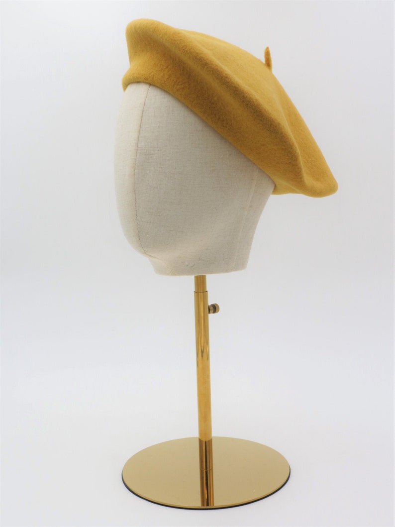 May include: A mustard yellow beret on a white mannequin head. The mannequin is on a gold stand with a round base.
