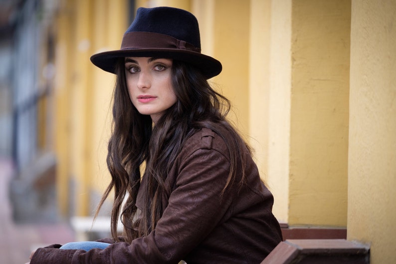 Op de afbeelding: Een vrouw met lang donker haar die een bruin su&egrave;de jack en een zwarte fedora met een bruine band draagt.